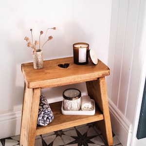 May include: A wooden step stool with a heart-shaped detail, holding a lit candle, a small vase with dried flowers, and a tray with cotton buds and a rolled towel. The stool is on a patterned tile floor. The candle is in a glass jar.