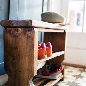K&ouml;nnte beinhalten: Ein Schuhregal aus Holz mit zwei Ablagen. Das Regal ist dunkelbraun gebeizt und hat einen rustikalen Look. Auf den Ablagen befinden sich Schuhe, darunter ein Paar rot-wei&szlig;e Sneaker und ein Paar pink-orangefarbene Sneaker.