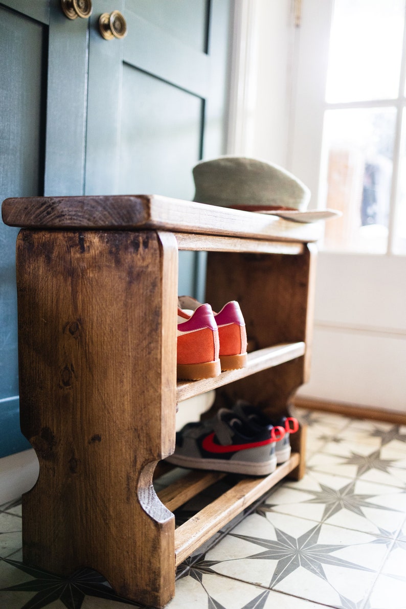 K&ouml;nnte beinhalten: Ein Schuhregal aus Holz mit zwei Ablagen. Das Regal ist dunkelbraun gebeizt und hat einen rustikalen Look. Auf den Ablagen befinden sich Schuhe, darunter ein Paar rot-wei&szlig;e Sneaker und ein Paar pink-orangefarbene Sneaker.