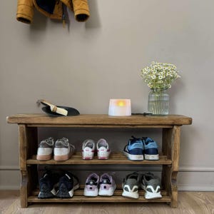 May include: A rustic, wooden shoe rack with two shelves, holding various sneakers. A small vase of white flowers, a candle, and a dustpan with a brush sit on top. A yellow jacket hangs above.