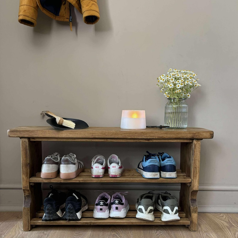 May include: A rustic, wooden shoe rack with two shelves, holding various sneakers. A small vase of white flowers, a candle, and a dustpan with a brush sit on top. A yellow jacket hangs above.