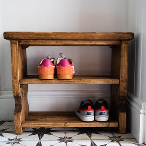 K&ouml;nnte beinhalten: Ein Schuhregal aus Holz mit zwei Ablagen. In der oberen Ablage stehen zwei Paar Sneaker, ein Paar ist orange und pink, das andere Paar ist schwarz und rot. In der unteren Ablage stehen zwei Paar Sneaker, beide sind schwarz und rot mit dem Schriftzug "NIKE" darauf.