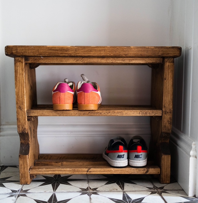 K&ouml;nnte beinhalten: Ein Schuhregal aus Holz mit zwei Ablagen. In der oberen Ablage stehen zwei Paar Sneaker, ein Paar ist orange und pink, das andere Paar ist schwarz und rot. In der unteren Ablage stehen zwei Paar Sneaker, beide sind schwarz und rot mit dem Schriftzug "NIKE" darauf.