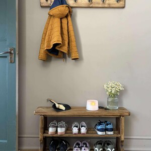 May include: A rustic wooden entryway bench with two shelves, holding various pairs of shoes. Above, a wooden coat rack with metal hooks supports a mustard-yellow quilted jacket with a blue hood. A candle and flowers sit on the bench.