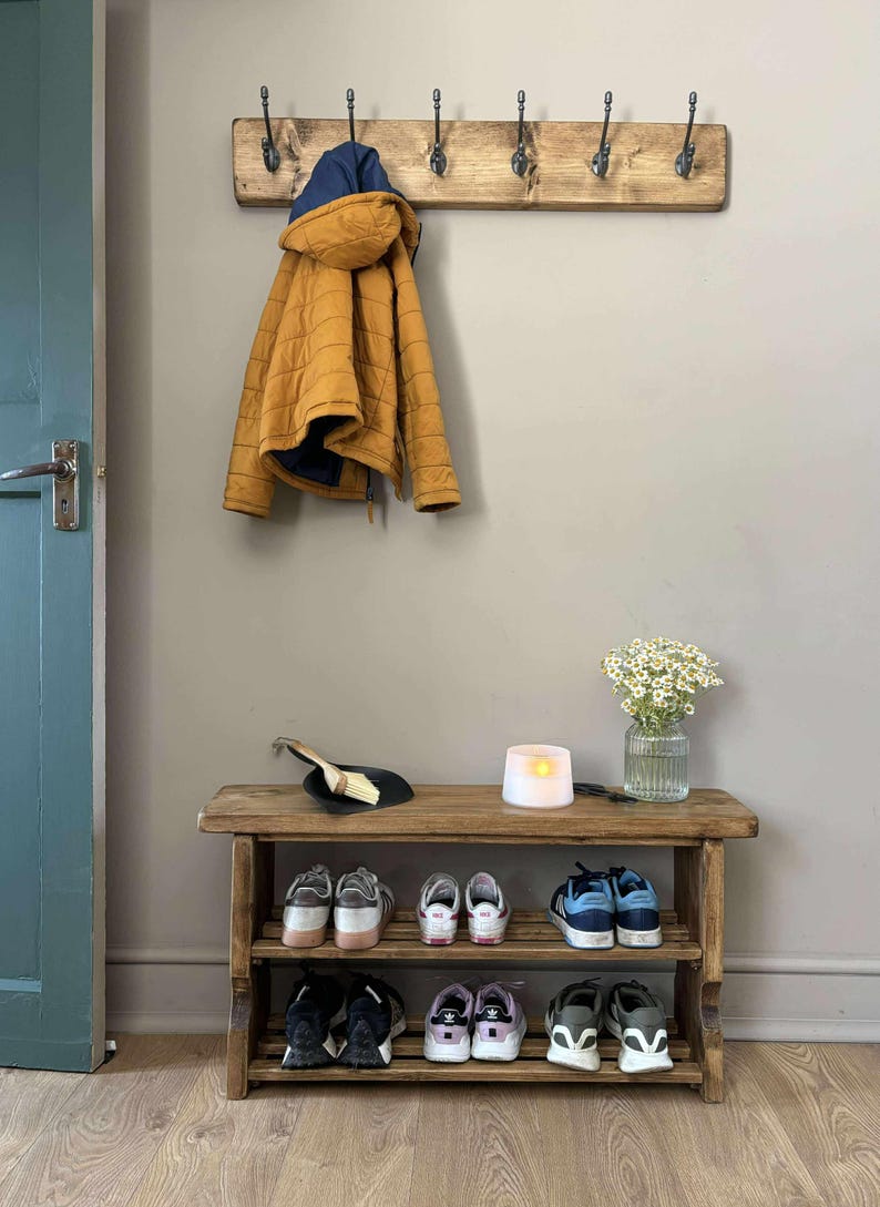 May include: A rustic wooden entryway bench with two shelves, holding various pairs of shoes. Above, a wooden coat rack with metal hooks supports a mustard-yellow quilted jacket with a blue hood. A candle and flowers sit on the bench.