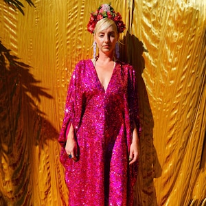 Könnte beinhalten: Eine Frau trägt eine rosa paillettenbesetzte Kaftan mit einer Blumenkrone und großen Ohrringen. Sie steht vor einem goldenen Hintergrund.