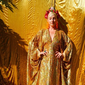 Könnte beinhalten: Eine Frau in einem goldenen Paillettenkleid mit einer Blumenkrone und großen Ohrringen. Das Kleid hat einen tiefen V-Ausschnitt und lange Ärmel. Die Frau steht vor einem goldenen Hintergrund.