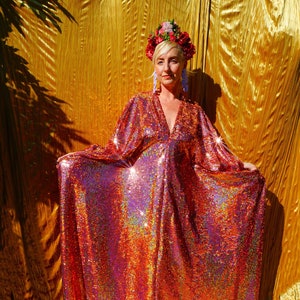 Könnte beinhalten: Eine Frau in einem langen, schimmernden, rosafarbenen Paillettenkleid mit einer Blumenkrone. Das Kleid hat einen tiefen V-Ausschnitt und lange Ärmel. Die Frau steht vor einem goldenen Hintergrund.