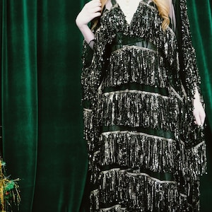 Puede incluir: Un vestido negro con lentejuelas y detalles de flecos en capas. El vestido tiene un escote en V y se combina con una capa transparente a juego. El conjunto se presenta sobre una cortina de terciopelo verde oscuro, creando un efecto dram&aacute;tico. El vestido es adecuado para ocasiones especiales.