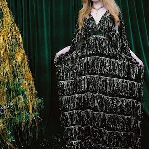 Puede incluir: Un vestido negro con lentejuelas y flecos en capas. El vestido tiene un escote en V y se lleva con un cintur&oacute;n negro. La modelo lleva un collar dorado. El fondo es una cortina verde oscuro.