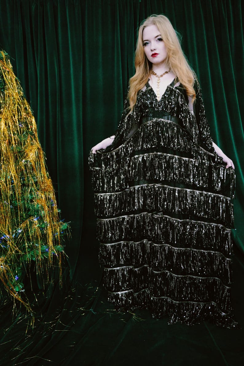 Puede incluir: Un vestido negro con lentejuelas y flecos en capas. El vestido tiene un escote en V y se lleva con un cintur&oacute;n negro. La modelo lleva un collar dorado. El fondo es una cortina verde oscuro.