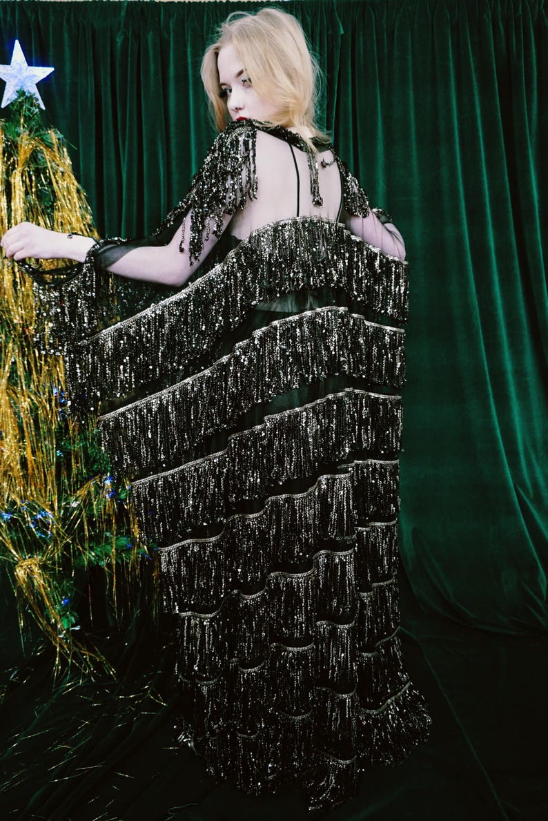 Puede incluir: Vestido negro con capas de flecos plateados. El vestido tiene la espalda y las mangas transparentes, y se lleva delante de una cortina de terciopelo verde. A la izquierda se ve un peque&ntilde;o &aacute;rbol de Navidad.