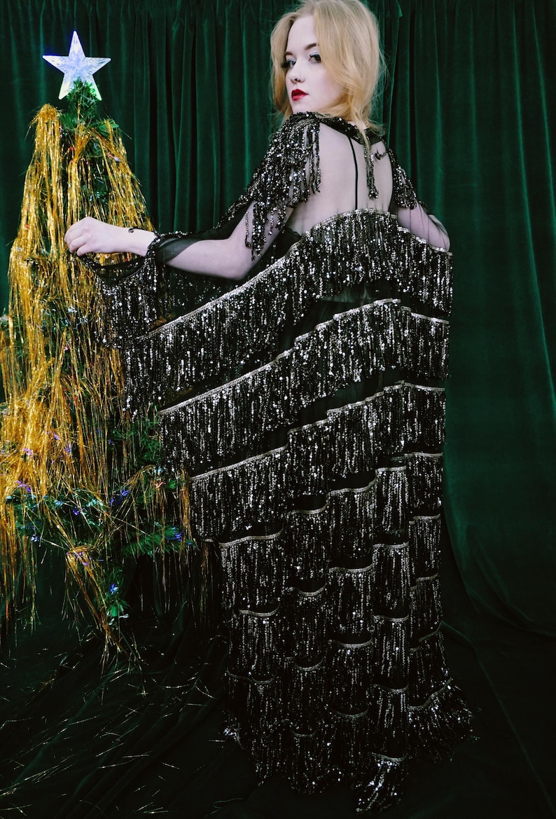 Puede incluir: Vestido negro largo con filas escalonadas de flecos plateados. El vestido tiene la espalda y las mangas transparentes. Un peque&ntilde;o &aacute;rbol de Navidad decorado con una estrella se encuentra en el fondo.