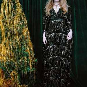 Puede incluir: Una escena festiva con un &aacute;rbol de Navidad adornado con oropel dorado y una estrella. Una mujer con un vestido negro con lentejuelas y flecos est&aacute; cerca. El fondo es una cortina verde oscuro.
