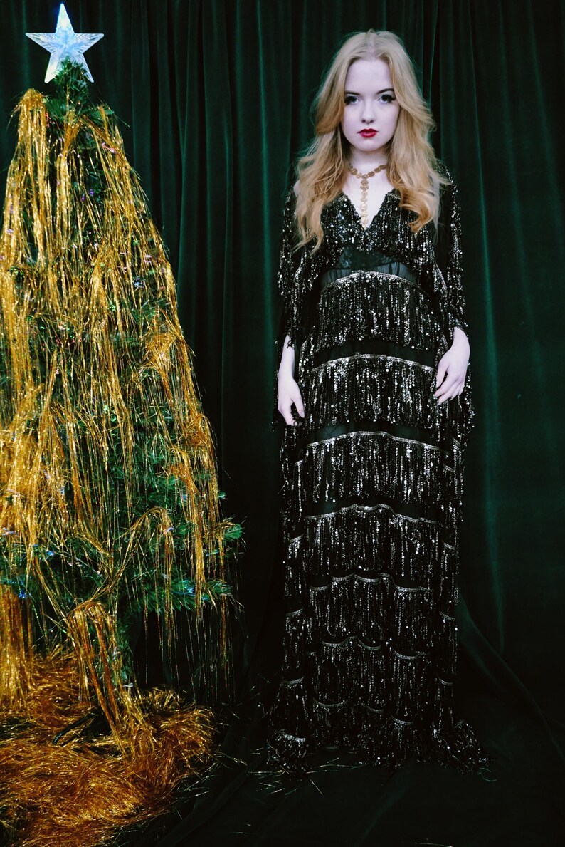 Puede incluir: Una escena festiva con un &aacute;rbol de Navidad adornado con oropel dorado y una estrella. Una mujer con un vestido negro con lentejuelas y flecos est&aacute; cerca. El fondo es una cortina verde oscuro.