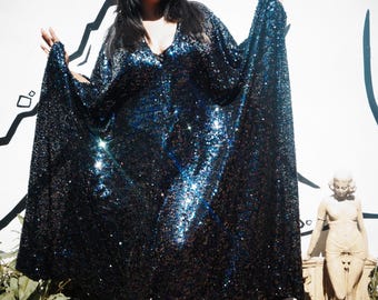 May include: A dark blue sequined dress with a V-neck and wide sleeves. The dress has a shimmering effect with blue highlights. The person is wearing a matching headband and silver clogs. A statue is to the right.