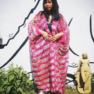 Könnte beinhalten: Ein leuchtend pinkfarbener, paillettenbesetzter Kimono-Stil-Bademantel mit goldenen Akzenten. Der Bademantel wird über einem schwarzen Oberteil getragen und mit silbernen Schuhen kombiniert. Ein passendes pinkfarbenes Stirnband vervollständigt den Look.