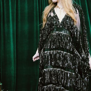 Puede incluir: Vestido negro con lentejuelas, cuello en V y flecos. El vestido tiene m&uacute;ltiples capas de flecos y se combina con una capa transparente. El fondo es una cortina verde oscuro.