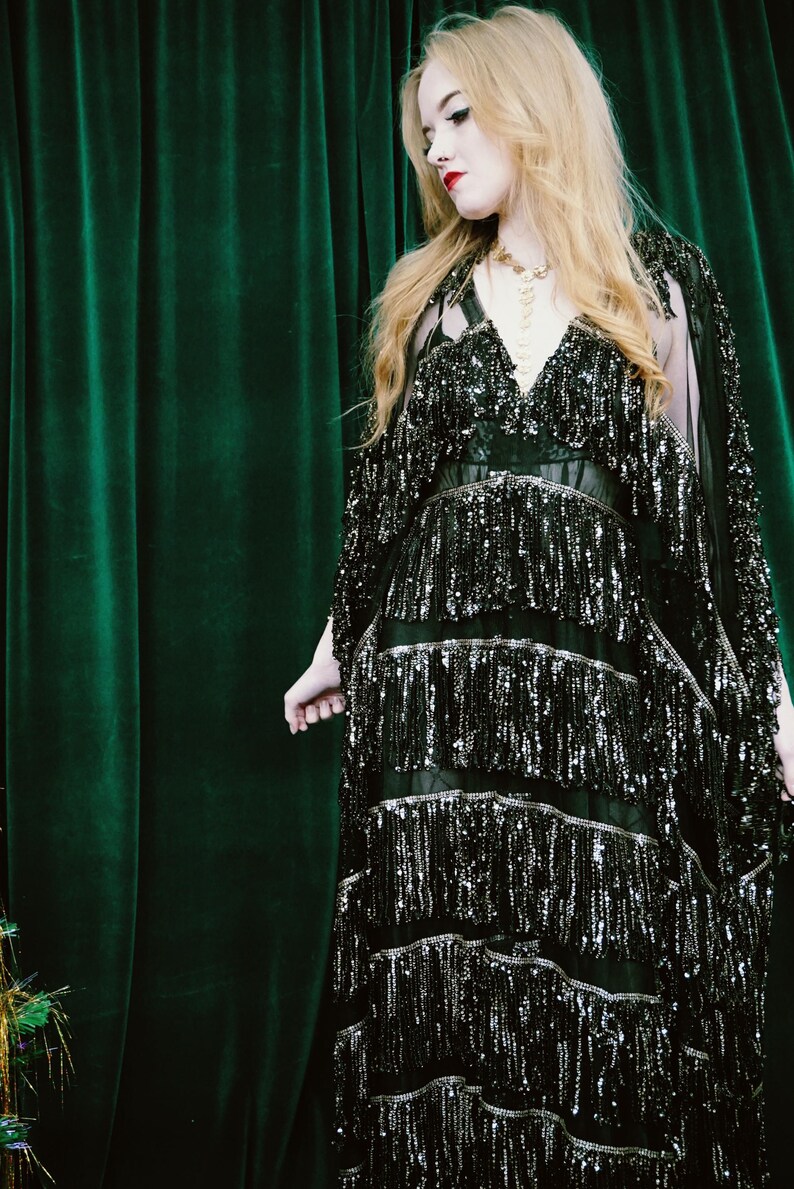 Puede incluir: Vestido negro con lentejuelas, cuello en V y flecos. El vestido tiene m&uacute;ltiples capas de flecos y se combina con una capa transparente. El fondo es una cortina verde oscuro.