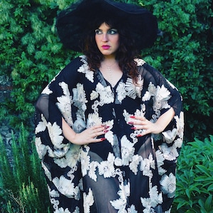 May include: A sheer black kaftan featuring an intricate white floral embroidery pattern, a deep V-neckline, and wide flowing sleeves. The garment is paired with a wide-brimmed black hat, creating a cohesive look with a dark, semi-transparent fabric.