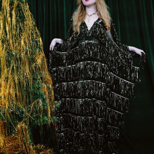 Puede incluir: Una persona con un vestido negro con flecos, de pie junto a un &aacute;rbol de Navidad adornado con oropel dorado y una estrella. El vestido tiene cuello en V y mangas largas. El fondo es una cortina verde oscuro.