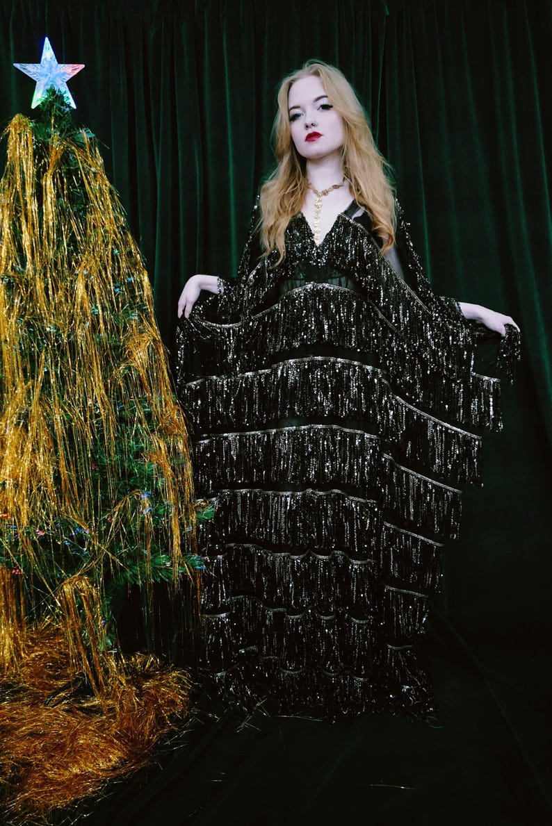 Puede incluir: Una persona con un vestido negro con flecos, de pie junto a un &aacute;rbol de Navidad adornado con oropel dorado y una estrella. El vestido tiene cuello en V y mangas largas. El fondo es una cortina verde oscuro.