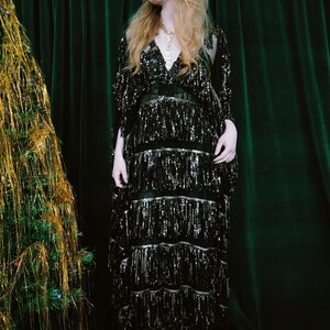 Puede incluir: Un vestido negro con lentejuelas y flecos. El vestido tiene cuello en V y mangas largas. La modelo lleva un collar dorado. El fondo es una cortina verde oscuro.