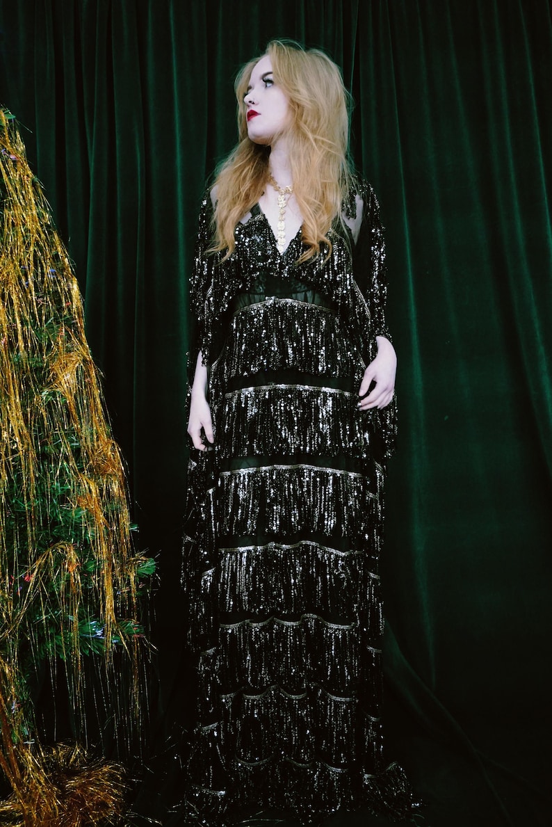 Puede incluir: Un vestido negro con lentejuelas y flecos. El vestido tiene cuello en V y mangas largas. La modelo lleva un collar dorado. El fondo es una cortina verde oscuro.