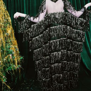 Puede incluir: Vestido negro con lentejuelas y flecos en capas. El vestido tiene mangas largas con adornos de lentejuelas y una espalda transparente. El fondo es una cortina de terciopelo verde oscuro y un &aacute;rbol de Navidad de oropel dorado.