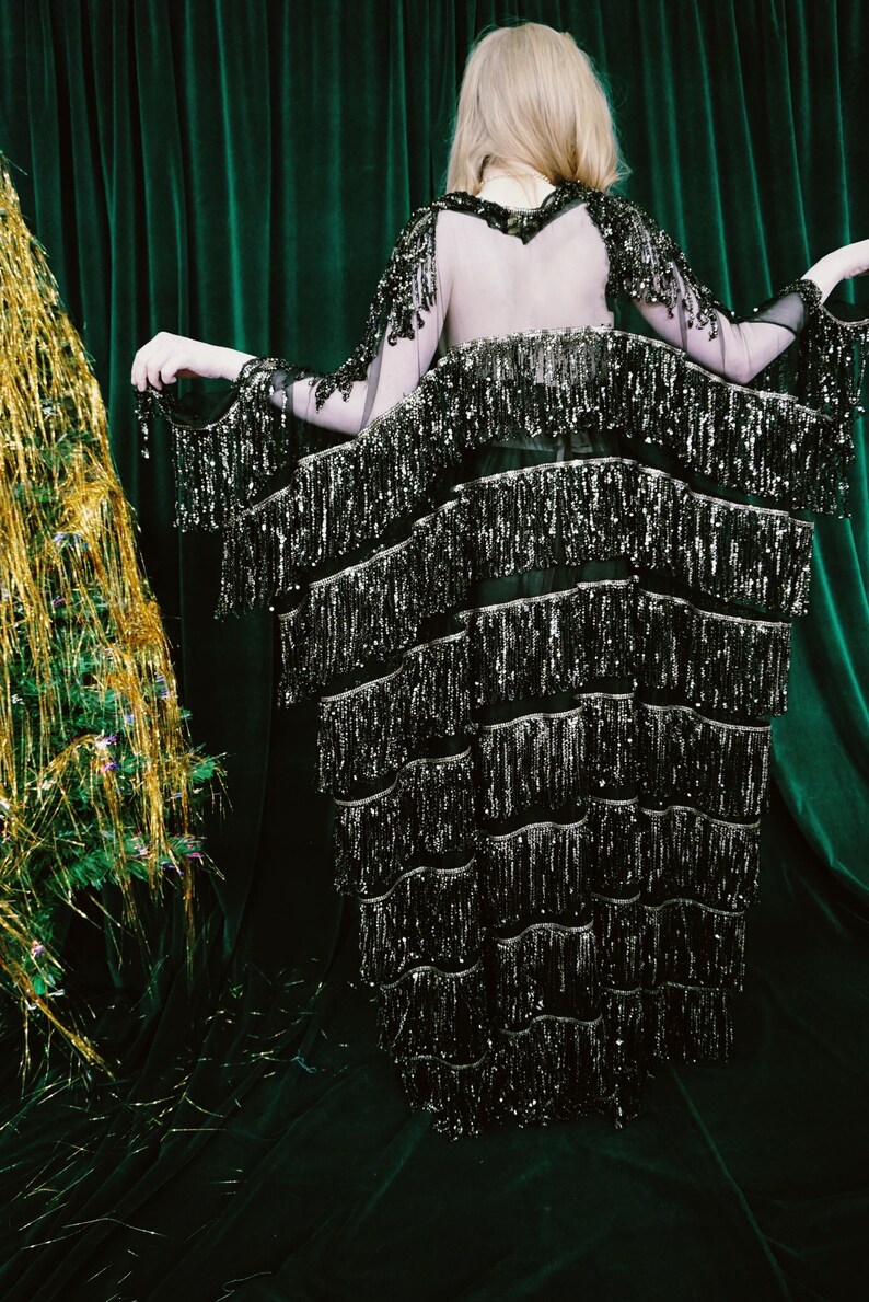 Puede incluir: Vestido negro con lentejuelas y flecos en capas. El vestido tiene mangas largas con adornos de lentejuelas y una espalda transparente. El fondo es una cortina de terciopelo verde oscuro y un &aacute;rbol de Navidad de oropel dorado.