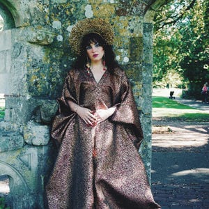 May include: A floor-length, wide-sleeved kaftan dress in a metallic bronze and dark purple fabric with a textured, crackled pattern. The garment features a V-neckline and a voluminous silhouette, paired with a large, circular, gold-toned halo headpiece.
