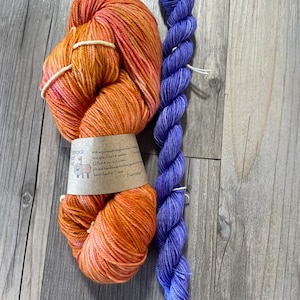 May include: Two skeins of yarn, one orange and one blue, rest on a wooden surface. The orange yarn is a large, loosely wound ball with a label. The blue yarn is a tightly wound, long skein. The label reads "Lakeside Fiber Co." and "Peach Mango."