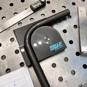 May include: A metal tube bent into a curve, positioned on a metal work surface. A digital angle finder and ruler are nearby. A black tool with the text "MA.LE HABT WORKS" is also present. A blueprint with the number 25 is visible.