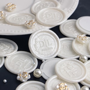May include: White wax seals with the initials "D|L" and the date "05.04.2023" embossed in gold. The seals are arranged on a black surface with white pearls and decorative flowers.
