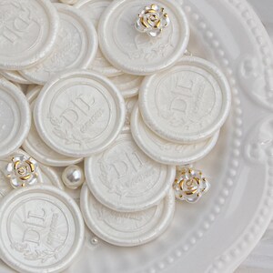 May include: A close-up of a white plate with multiple white wax seals with the initials "DIL" and the word "COUTURE" embossed on them. There are also small white pearls and gold-colored rose embellishments scattered on the plate.