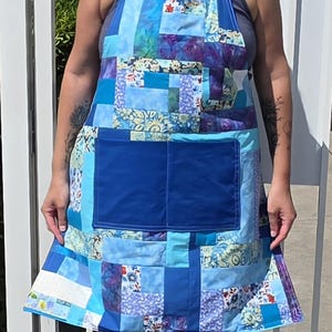 May include: A patchwork apron featuring a variety of blue, light blue, and white fabric squares with floral and abstract patterns. The apron includes a large, solid dark blue rectangular pocket centered on the front and blue neck straps.