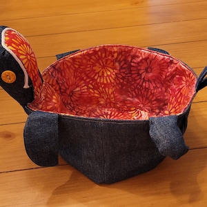 May include: A denim turtle-shaped basket with a red and orange floral fabric lining. The basket has two handles and a button on the head.