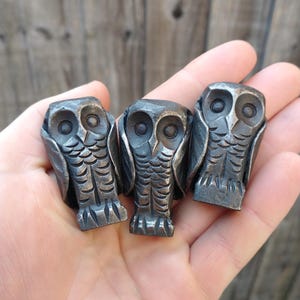 May include: Three handcrafted metal owl figurines. Each owl has detailed feather and eye features. The owls are a dark, metallic color and are held in a hand against a wooden background. The figurines are approximately 2 inches tall.