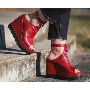 Peut inclure: Sandales compensées en cuir rouge avec un bout ouvert et des brides de cheville. Les sandales ont une semelle noire et un talon compensé haut. Les chaussures sont portées avec un jean en denim foncé.