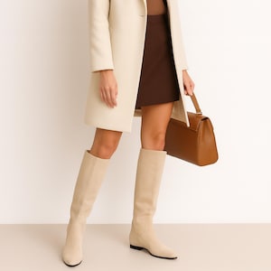 Puede incluir: Un abrigo color crema y botas hasta la rodilla se combinan con una falda marrón y un bolso de mano de cuero marrón. Las botas tienen suela plana y una textura similar a la gamuza. El abrigo está abotonado y el bolso tiene cierre de solapa.