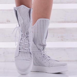 May include: White perforated leather ankle boots with laces. The boots have a thick white sole and are shown on a white wooden background.