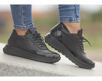 May include: Black platform sneakers with a glossy patent leather accent on the toe and heel. The sneakers have a thick, textured sole and are laced up. The shoes are worn with blue jeans.