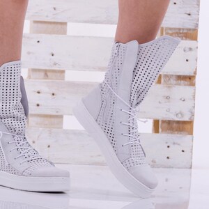 May include: White perforated leather ankle boots with laces. The boots have a thick white sole and are shown on a model's feet.