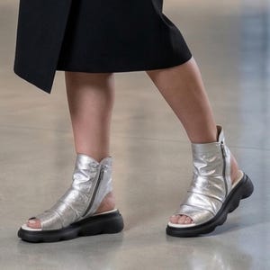 以下が含まれることがあります： 黒いソールとサイドジッパーが付いた、シルバーのオープントゥサンダル。 サンダルはギャザーのデザインで、白いインソールが付いています。 黒いスカートを履いた人が履いています。