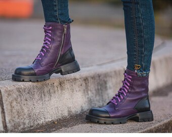 May include: Purple leather combat boots with black detailing, a zip, and purple laces. The boots feature a chunky sole and are worn with blue jeans. These boots are a fashionable and edgy footwear option.