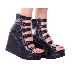 May include: Black platform wedge sandals with multiple straps and buckles. The open-toe shoes have a chunky, ridged sole and a zipper on the side. The sandals are made of a smooth, matte material.