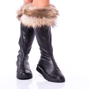 Könnte beinhalten: Schwarze, kniehohe Lederstiefel mit einem braunen Kunstfellbesatz. Die Stiefel haben eine spitze Zehenpartie und einen niedrigen Absatz.