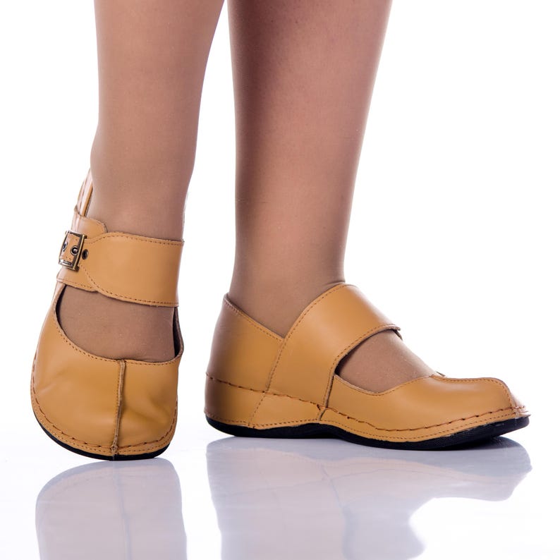 May include: A pair of tan leather shoes with a buckle strap and a closed toe. The shoes have a slightly raised heel.