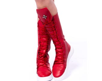 Puede incluir: Botas rojas de cuero hasta la rodilla con suelas y cordones blancos. Las botas tienen un detalle de cruz plateada en la parte superior.