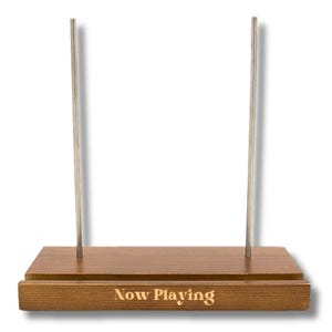 May include: A wooden record stand with two silver metal rods. The base is rectangular and has the words "Now Playing" engraved on the front. The wood has a warm, brown tone.